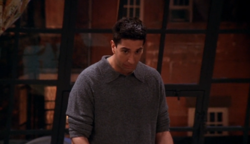 A man in a gray sweater standing in front of a window.