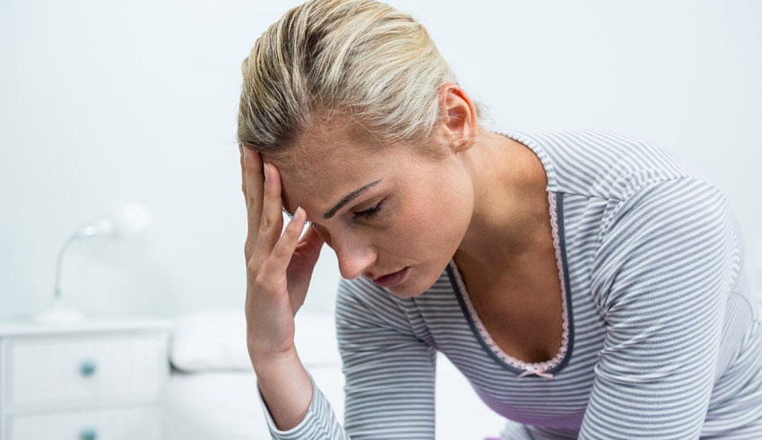 A woman is sitting on her bed with her hand on her head.