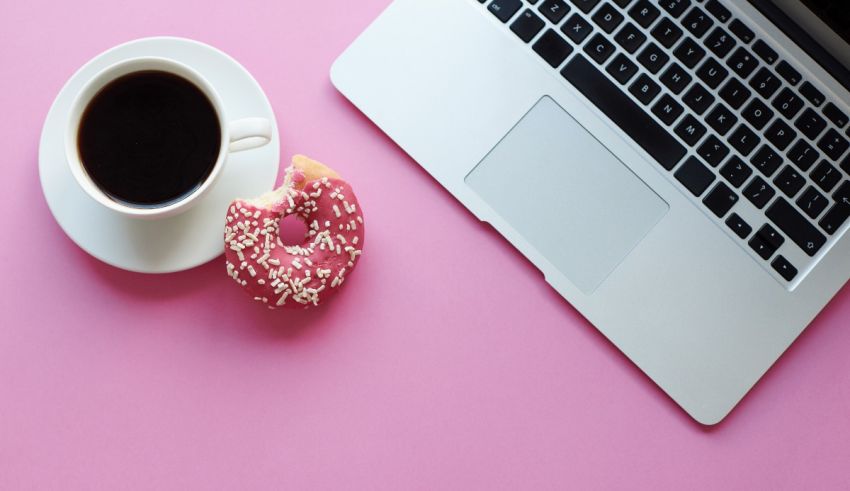 A cup of coffee and a donut next to a laptop on a pink background.