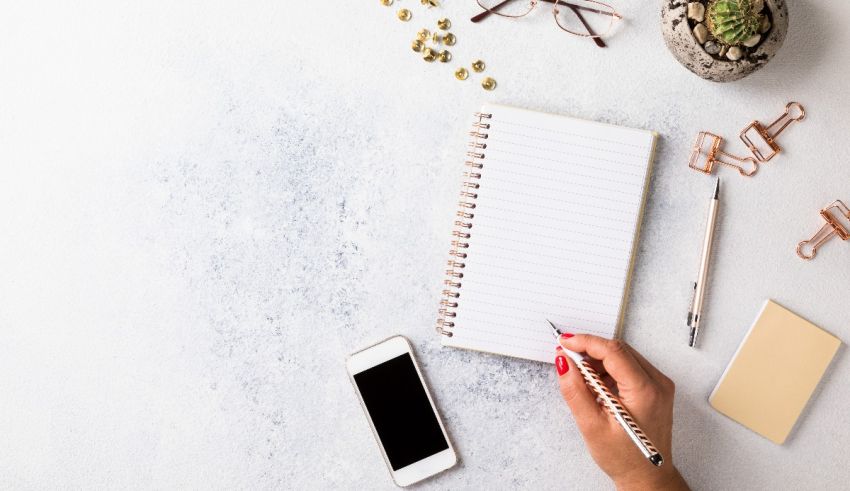 A woman's hand writing on a notepad next to a phone and a plant.