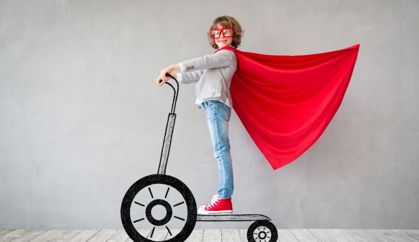 A boy wearing a red cape riding a scooter.