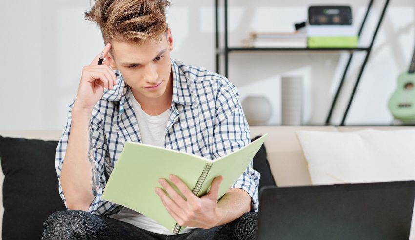 A young man sitting on a couch reading a book and using a laptop.