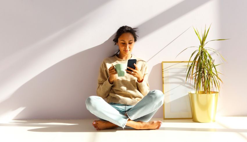 A woman sitting on the floor with her phone and a plant in front of her.