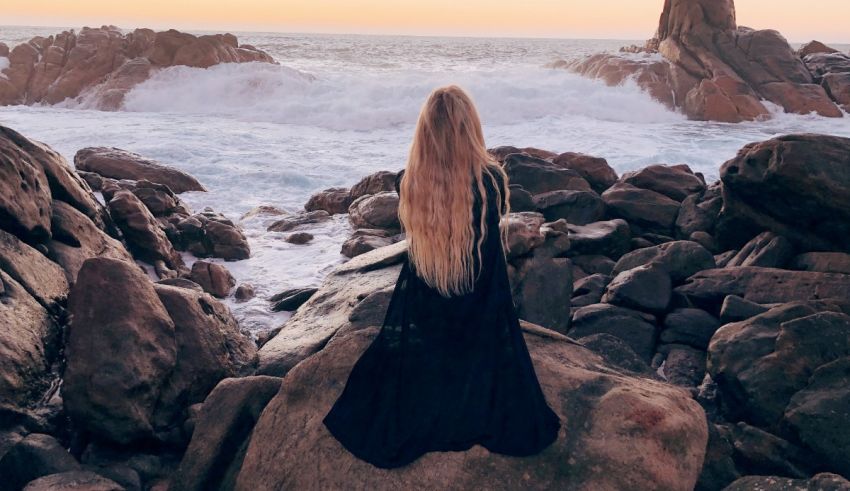 A woman with long hair standing on a rock looking at the ocean.