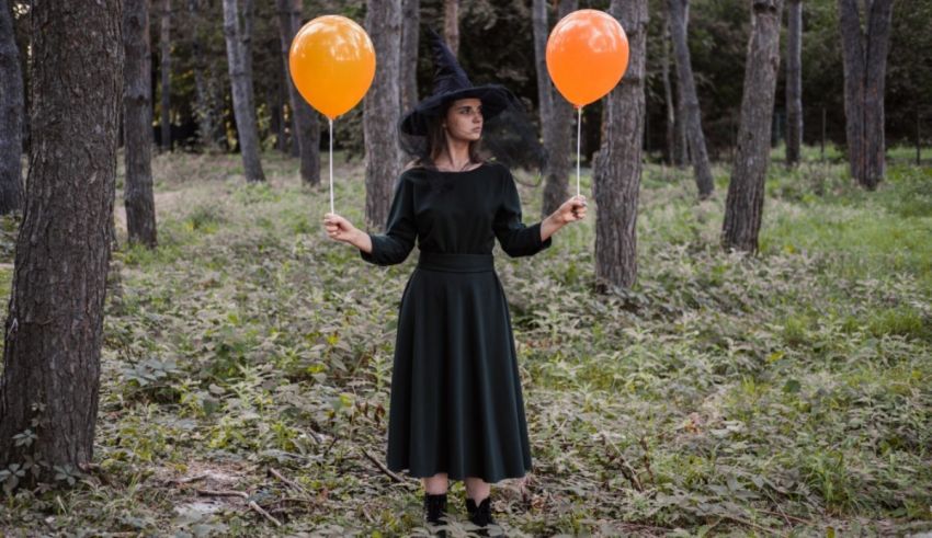 A woman in a witch hat holding orange balloons in the woods.