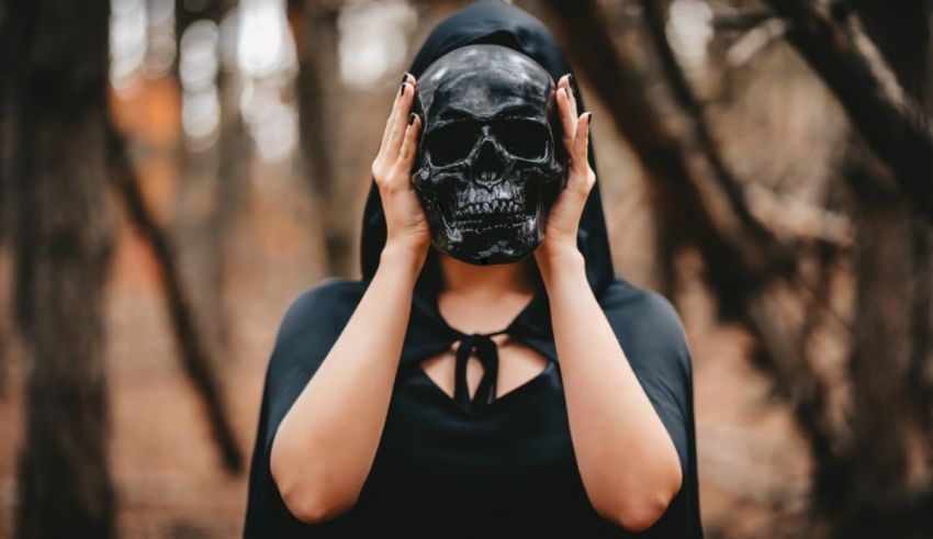 A woman wearing a black cloak and a skull mask in the woods.
