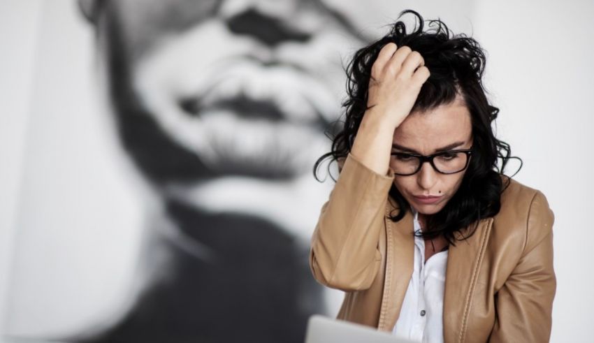 A woman with glasses is holding her head while working on a laptop.
