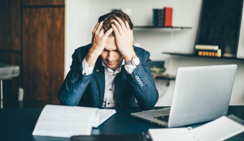 A man is holding his head while sitting at a desk with a laptop.