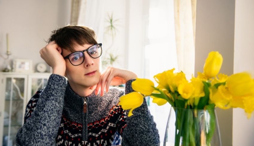 A young man in glasses sits at a table with a vase of daffodils.