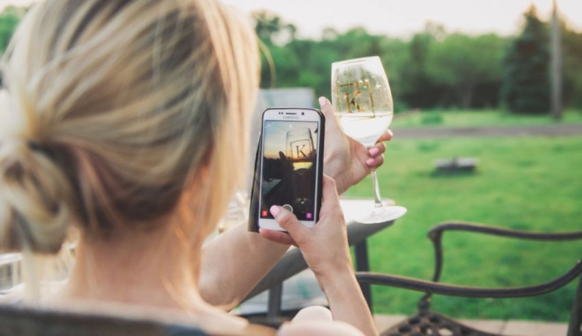 A woman holding a wine glass and taking a photo on her phone.