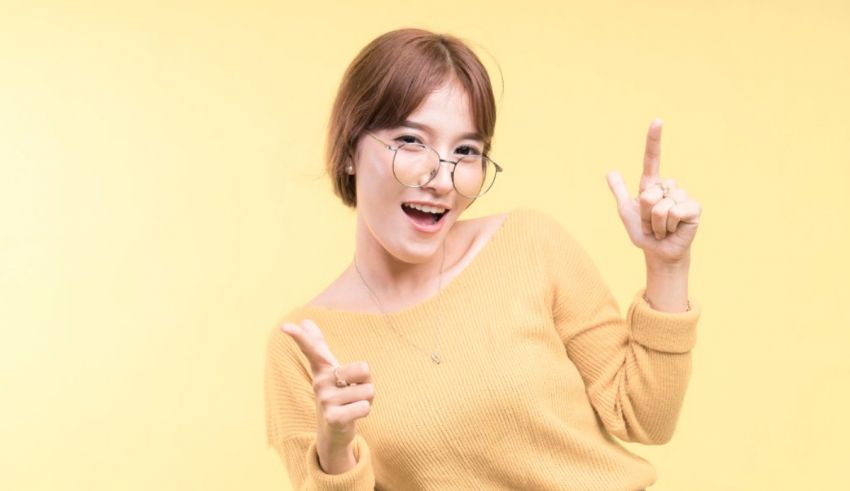 Asian woman with glasses pointing her finger at the camera on a yellow background.