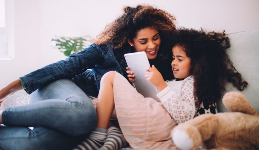 A mother and daughter are sitting on a couch looking at a tablet.