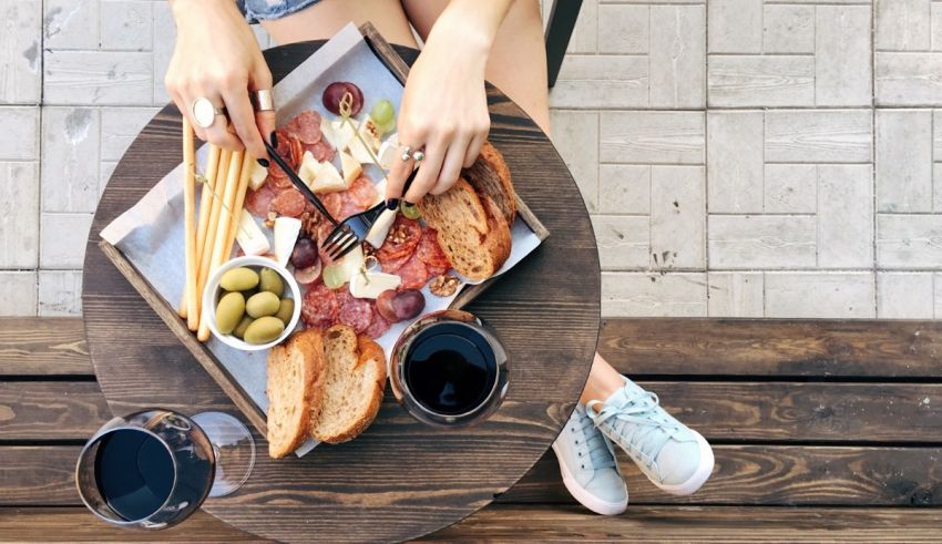 A woman is sitting on a wooden bench with a plate of food and wine.