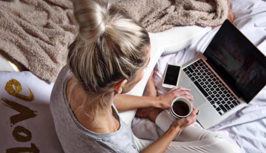 A woman working on her laptop in bed with a cup of coffee.