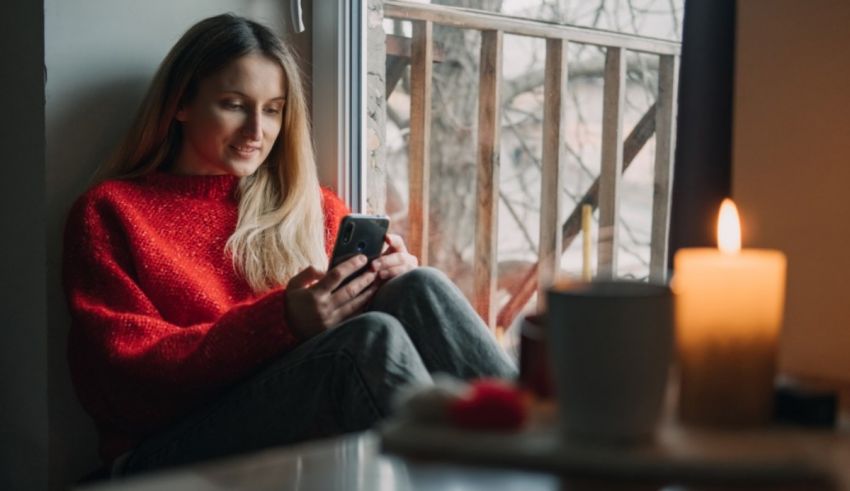 A woman is sitting in front of a window looking at her phone.