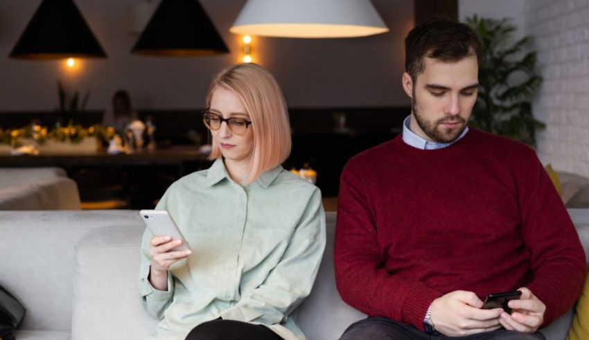 A man and woman sitting on a couch looking at their cell phones.