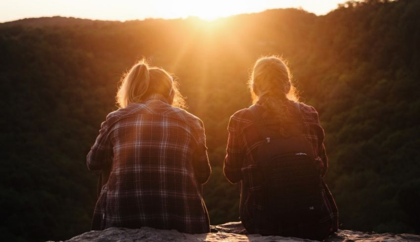 Two women sitting on top of a mountain looking at the sun.
