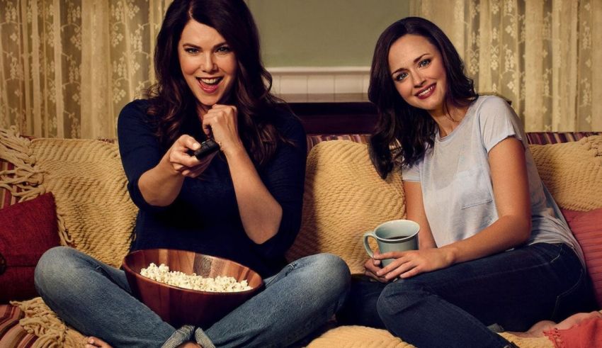 Two women sitting on a couch with popcorn and a remote.