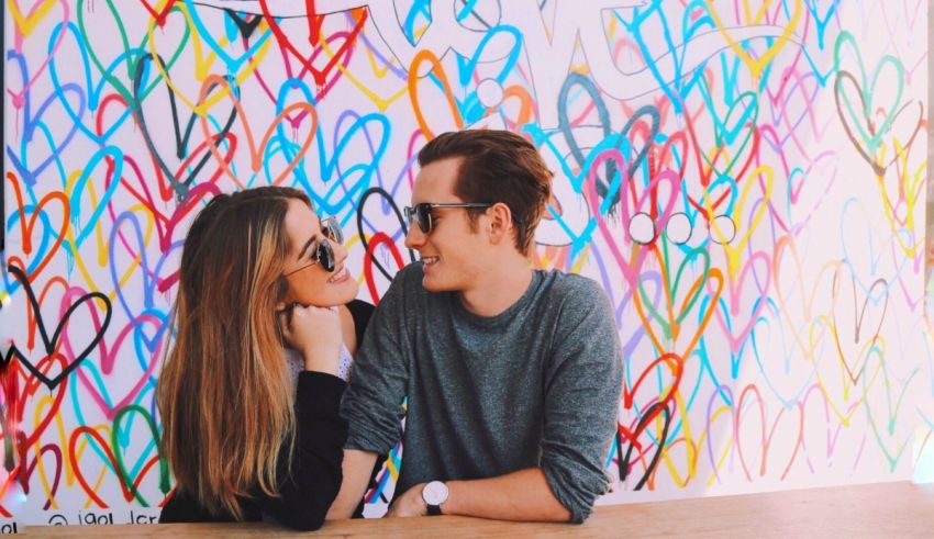 A man and woman sitting at a table with a wall of hearts.