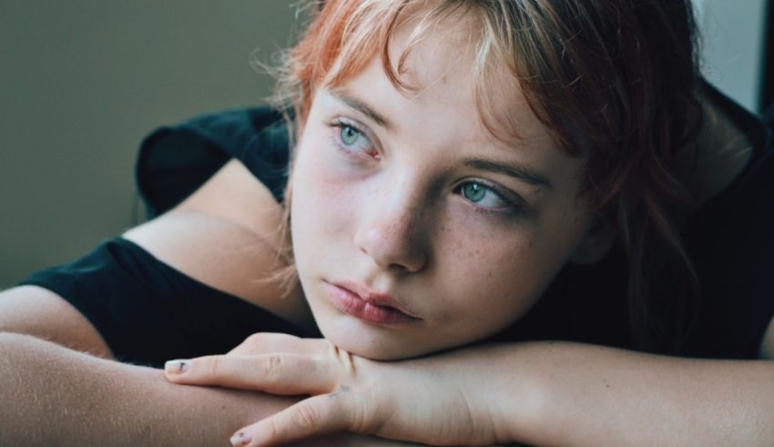 A young girl with red hair is leaning against a window.