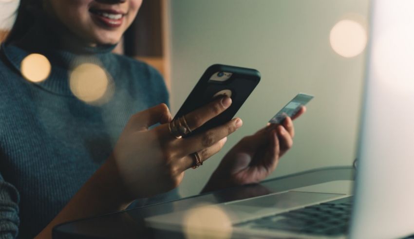A woman is holding a cell phone and a credit card in front of a laptop.