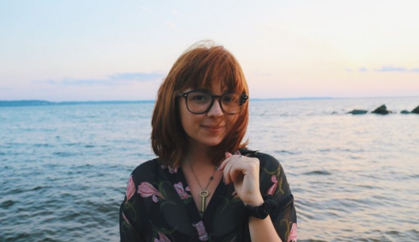 A woman with red hair and glasses standing in front of water.