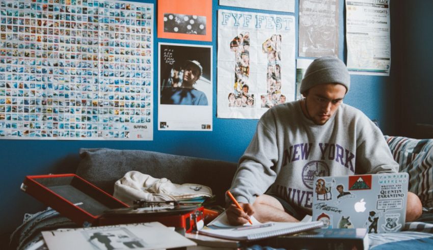 A man sitting on a bed with a laptop in front of a blue wall.