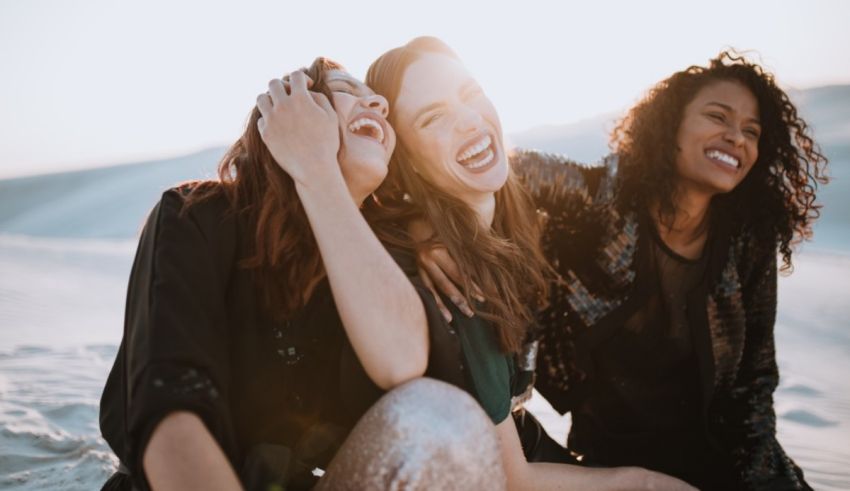 Three women laughing while sitting on a sand dune.
