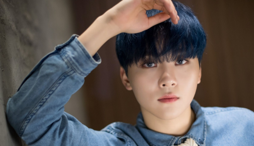 A young man with blue hair leaning against a wall.