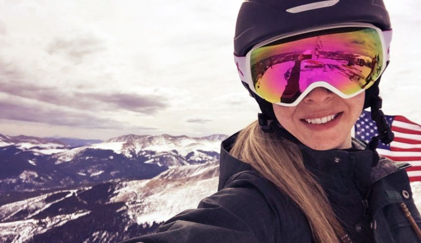 A woman wearing ski goggles and an american flag on top of a mountain.