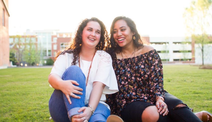 Two young women sitting on the grass in front of a building.