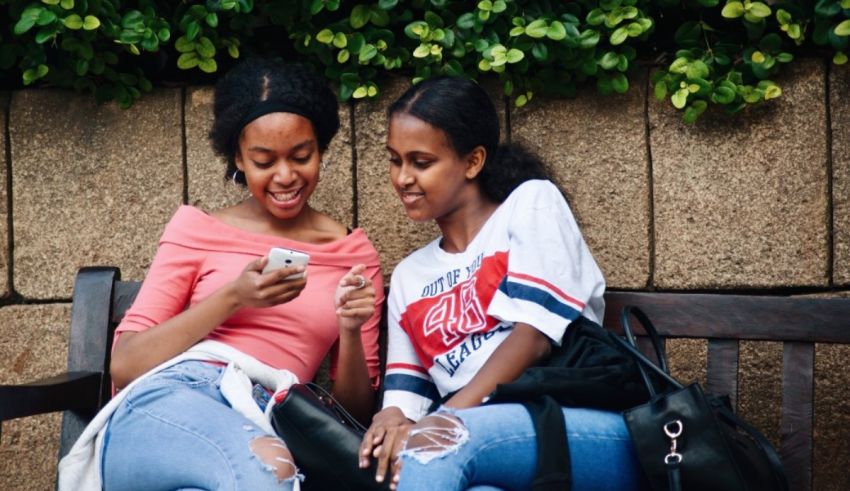 Two young women sitting on a bench looking at their cell phones.