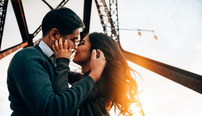 A couple kissing in front of a bridge at sunset.