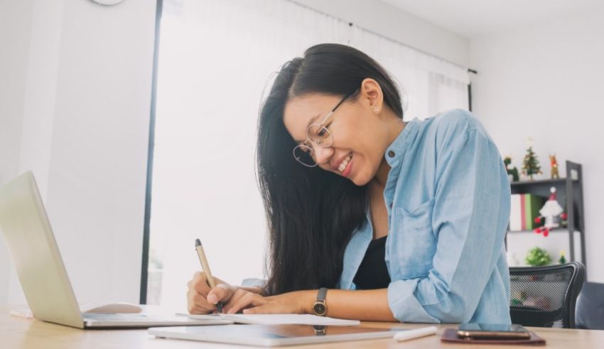A smiling asian woman writing on a laptop in her home office.