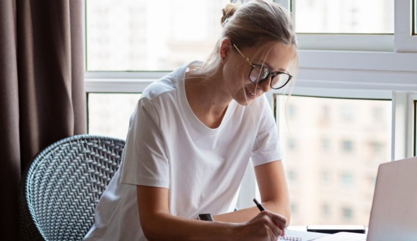 A woman writing on a laptop in front of a window.