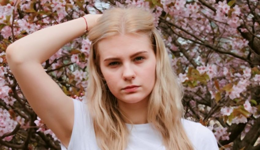 A young blonde woman in a white t-shirt posing in front of a blossoming tree.