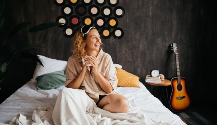 A young woman is sitting on a bed listening to music and drinking a cup of coffee.