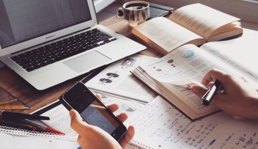 A person is using a cell phone while sitting at a desk with books and a laptop.