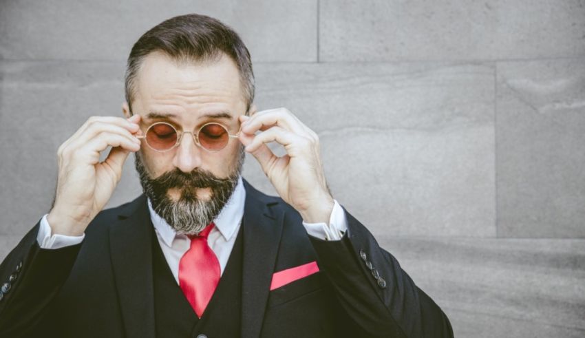 A bearded man in a suit and tie is looking at his glasses.