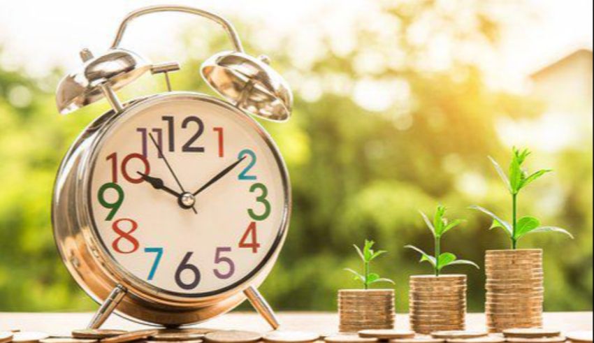 An alarm clock next to a stack of coins with a plant growing out of it.