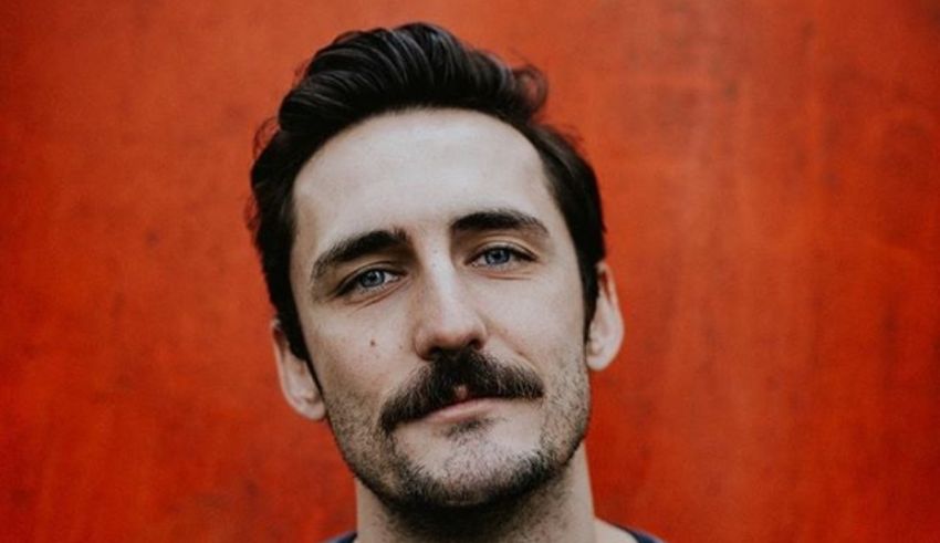 A man with a mustache in front of a red wall.