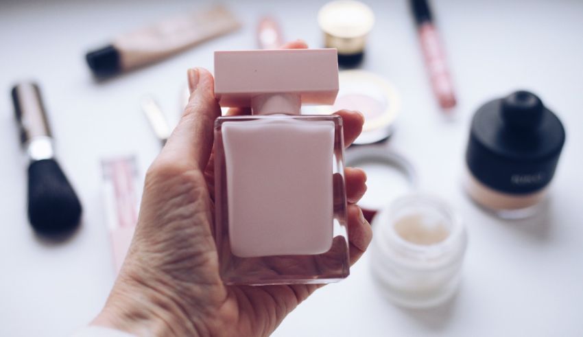 A hand holding a pink perfume bottle in front of cosmetics.