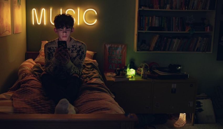 A young man sitting on a bed in a room with a neon music sign.