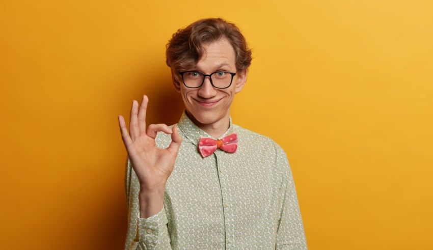 A young man with glasses and a bow tie showing the ok sign on a yellow background.
