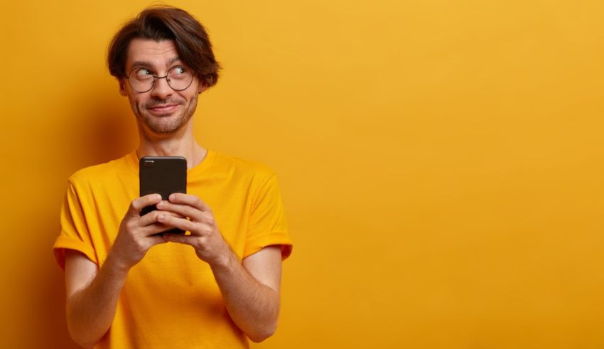 A man with glasses is holding a smart phone against a yellow background.