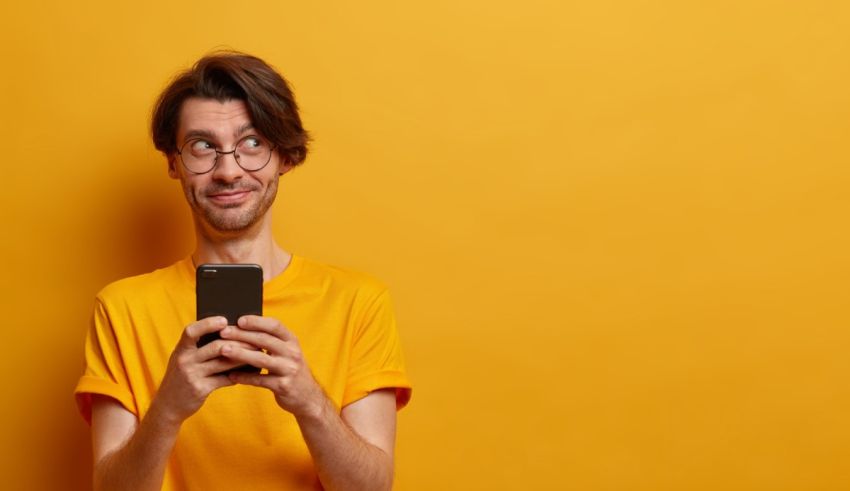 A young man with glasses is holding a smart phone against a yellow background.