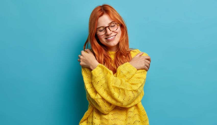 A young woman with red hair wearing glasses and a yellow sweater posing against a blue background.