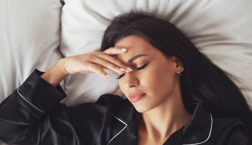 A woman in black pajamas laying in bed with her hand on her head.