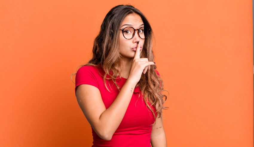 A woman wearing glasses is making a hush sign against an orange background.
