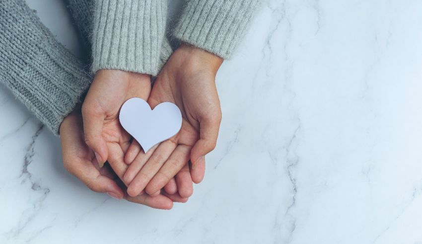 Two hands holding a heart shaped paper on a marble table.